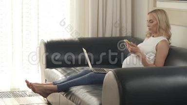 一个怀孕的金发女郎坐在沙发上，<strong>拿</strong>着<strong>手机</strong>和笔记本电脑的侧视图。 她<strong>拿</strong>着<strong>手机</strong>